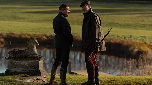 Tom hardy and pierce brosnan, dressed as a rich english country gentleman speak outside near a pond