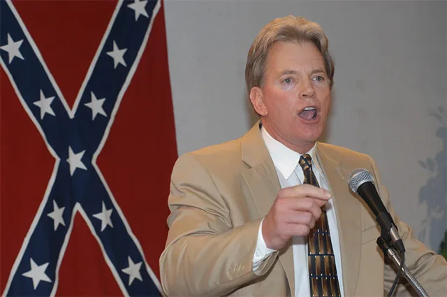 David Duke speaking at a podium with a Confederate flag in the background, wearing a light brown suit and tie, gesturing with his hand.