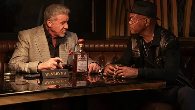 Promotional image of Sylvester Stallone and Samuel L. Jackson in "NOLA King" spinoff, sitting at a reserved table with a bottle of Montague Distillers whiskey, discussing in a dimly lit bar, highlighting their roles in the Tulsa King universe expansion by Taylor Sheridan.