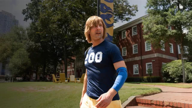 Glen Powell in Hulu's upcoming Chad Powers series, wearing a blue football jersey with the number 200, yellow shorts, and a blue arm sleeve, walking on a college campus with red brick buildings and green trees in the background.
