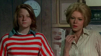 odie Foster and Barbara Harris in the 1976 Freaky Friday movie, standing in a vintage kitchen with a clock on the wall. Jodie Foster wears a red and white striped sweater, while Barbara Harris is dressed in a striped blouse, showcasing a classic mother-daughter body-swap comedy scene.