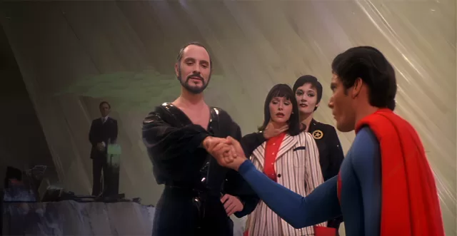 Terence Stamp as General Zod shaking hands with Christopher Reeve as Superman in a dramatic scene from Superman II, with Margot Kidder as Lois Lane looking concerned, Gene Hackman as Lex Luthor in the background, and Sarah Douglas as Ursa standing nearby, set against a futuristic backdrop.