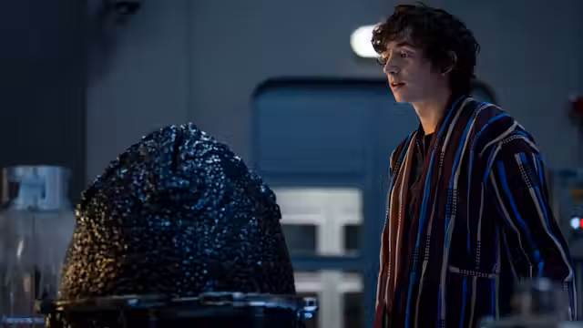 A young man in a striped robe stands in a dimly lit sci-fi laboratory from 'Alien Earth,' observing a large, dark, textured alien creature encased in a containment unit, with futuristic equipment and a blue-lit background.