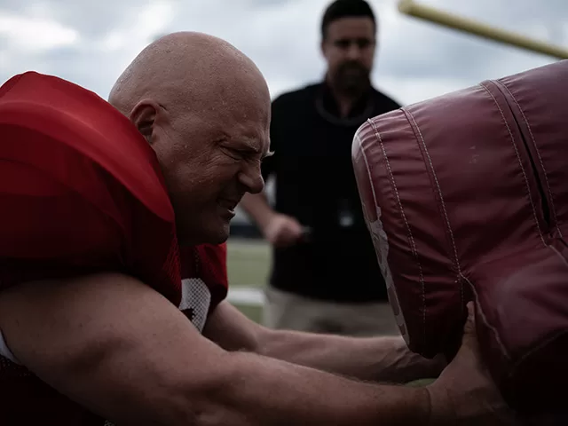 Michael Chiklis in The Senior movie, training intensely on the field, wearing a red Lobo football uniform, pushing against a tackling dummy with a coach in the background