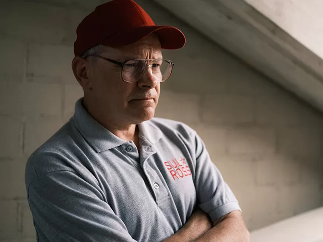 Rob Corddry as a coach in Angel Studios The Senior movie, wearing a red cap and gray polo shirt with arms crossed, standing in a stadium setting