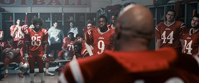 Michael Chiklis in The Senior movie, standing in a locker room with the Lobo football team, wearing a red jersey, surrounded by teammates in red uniforms.