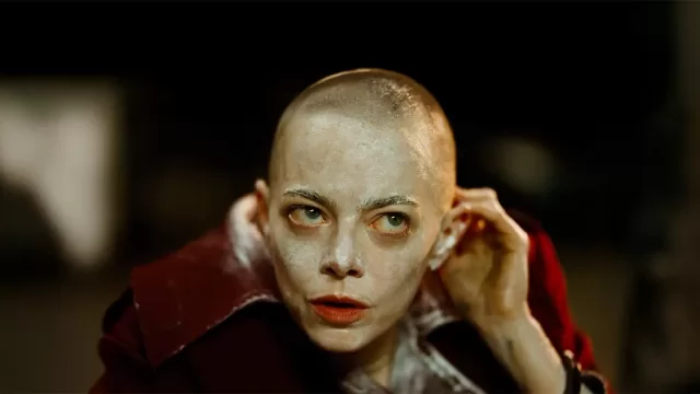 Emma Stone as Eun-Young in Bugonia: dramatic close-up portrait of the actress with shaved head, pale makeup, red lips, and intense green eyes, wearing a crimson coat collar in Yorgos Lanthimos' sci-fi thriller remake.