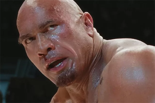 Dwayne "The Rock" Johnson as Mark Kerr in The Smashing Machine, showcasing an intense close-up of the muscular fighter with sweat glistening on his bald head and tattooed shoulder, captured mid-action in a gritty MMA ring scene