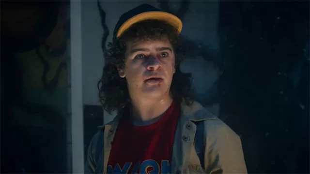 Dustin Henderson (Gaten Matarazzo) in Stranger Things season 5: classic curly hair, yellow cap, red MOM t-shirt, and denim jacket looking shocked in dramatic lighting – final season on Netflix – is Stranger Things 5 worth watching or ruined by woke agenda? Full conservative review and parental guide at WorthItOrWoke.com