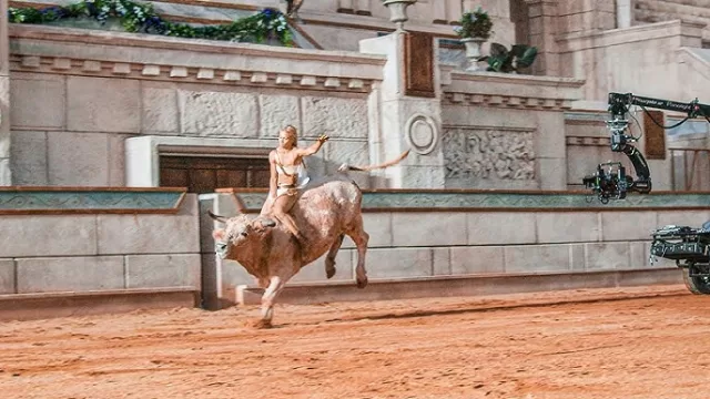 Rise of Merlin Daily Wire 2026 Charis bull ride scene: heroic figure in minimal garb atop raging bull in grand stone arena with columns, garlands, and film crane – Jeremy Boreing directed Arthurian origin – biblical values fantasy, anti-woke alternative to Hollywood, violence warnings, and Christian family rating on WorthItOrWoke.com
