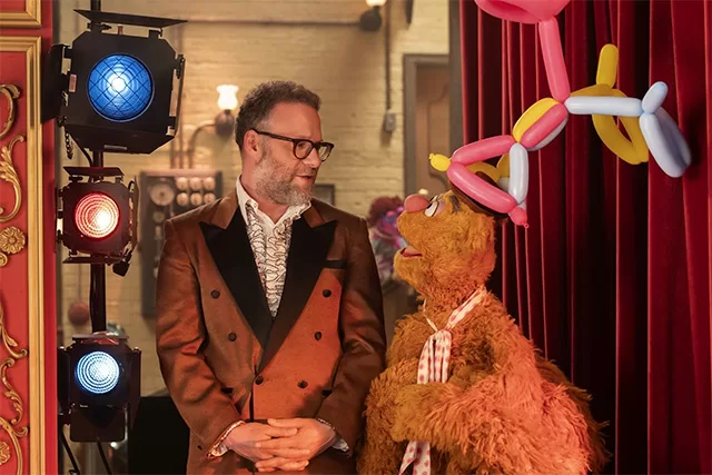 Seth Rogen Fozzie Bear Muppet Show 2026 scene: bearded Seth Rogen in tuxedo standing beside Fozzie with pink/yellow balloon hat on red theater curtain – Disney+ special event – significant sexual innuendos, crude humor complaints, woke agenda check, content warnings, and Christian family rating on WorthItOrWoke.com